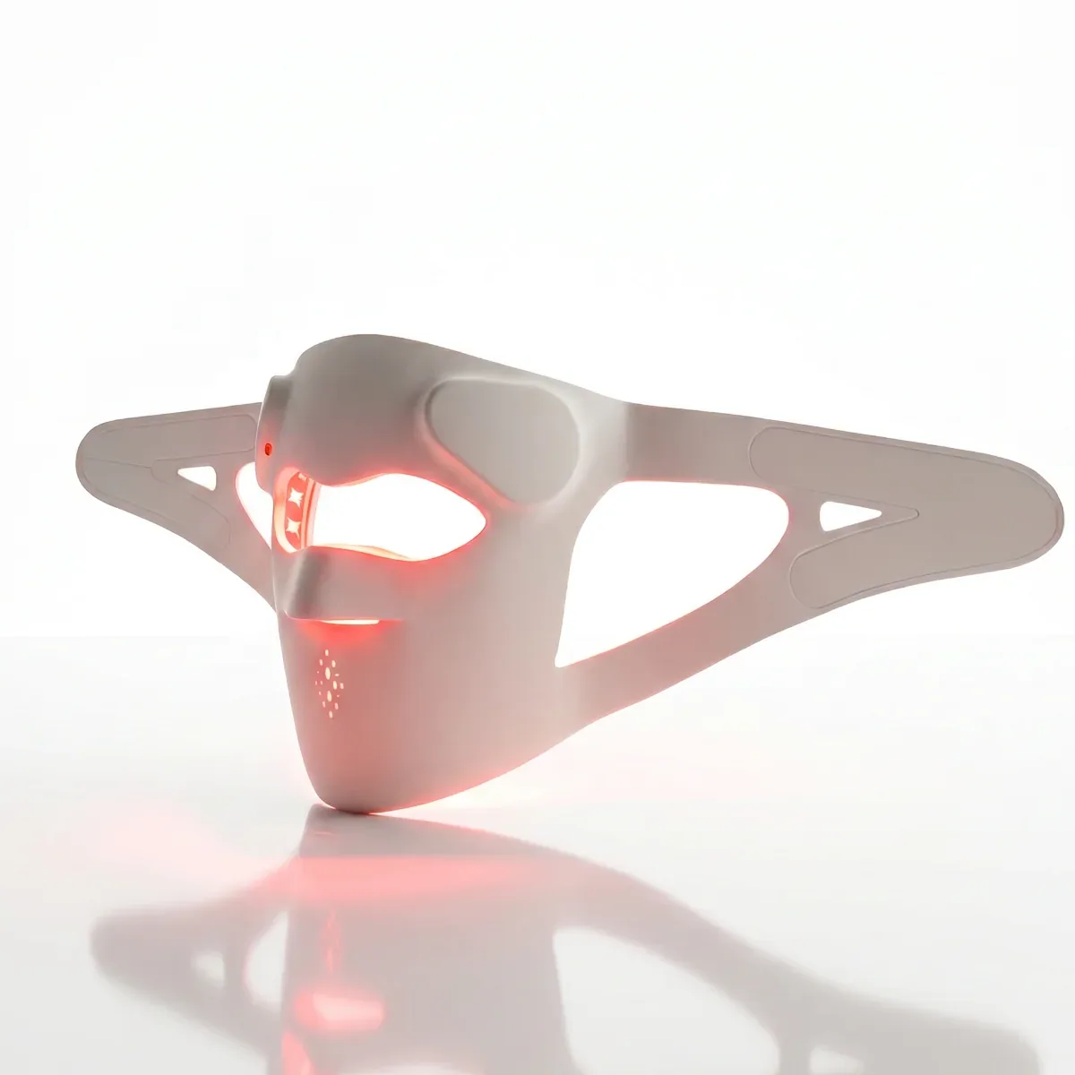 Dr. Renú SpectraLift Advanced LED face mask from a three-quarter angle, red light glowing through the eye and mouth openings, with its reflection on a white surface